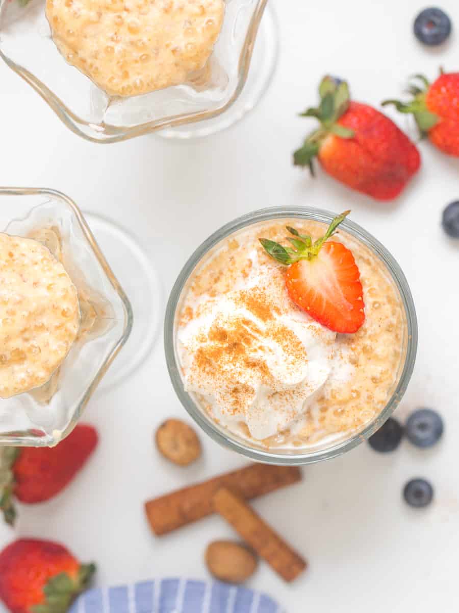 Tapioca pudding in small serving bowls topped with fresh whipped cream and strawberries with berries and cinnamon sticks in background.