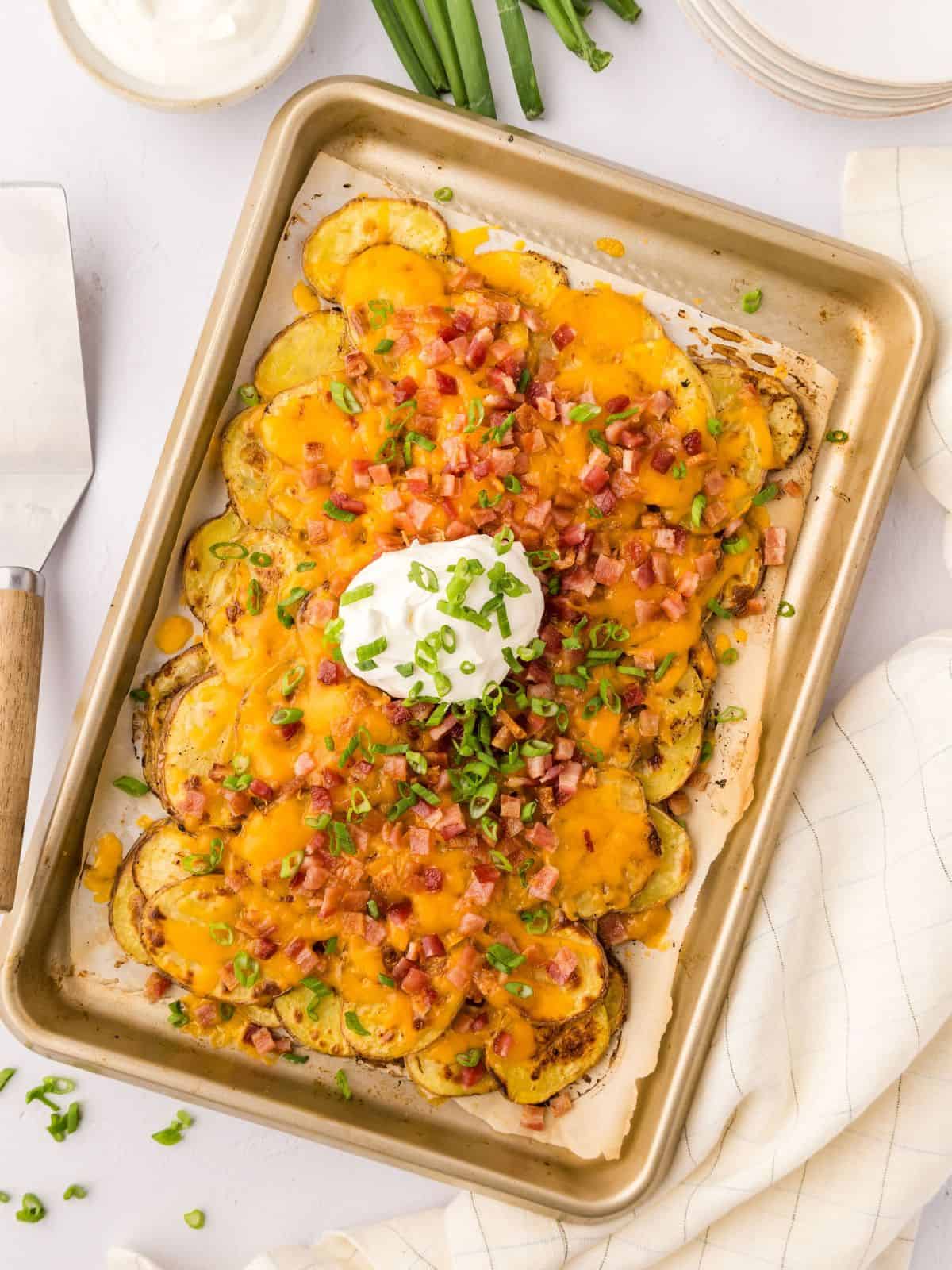 Loaded Irish Potato Nachos made with crispy baked potato slices topped with cheese and bacon on sheet pan topped with green onions and sour cream.