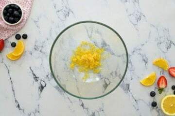 Lemon zest and granulated sugar combined in large mixing bowl with fresh lemon slices off to side.