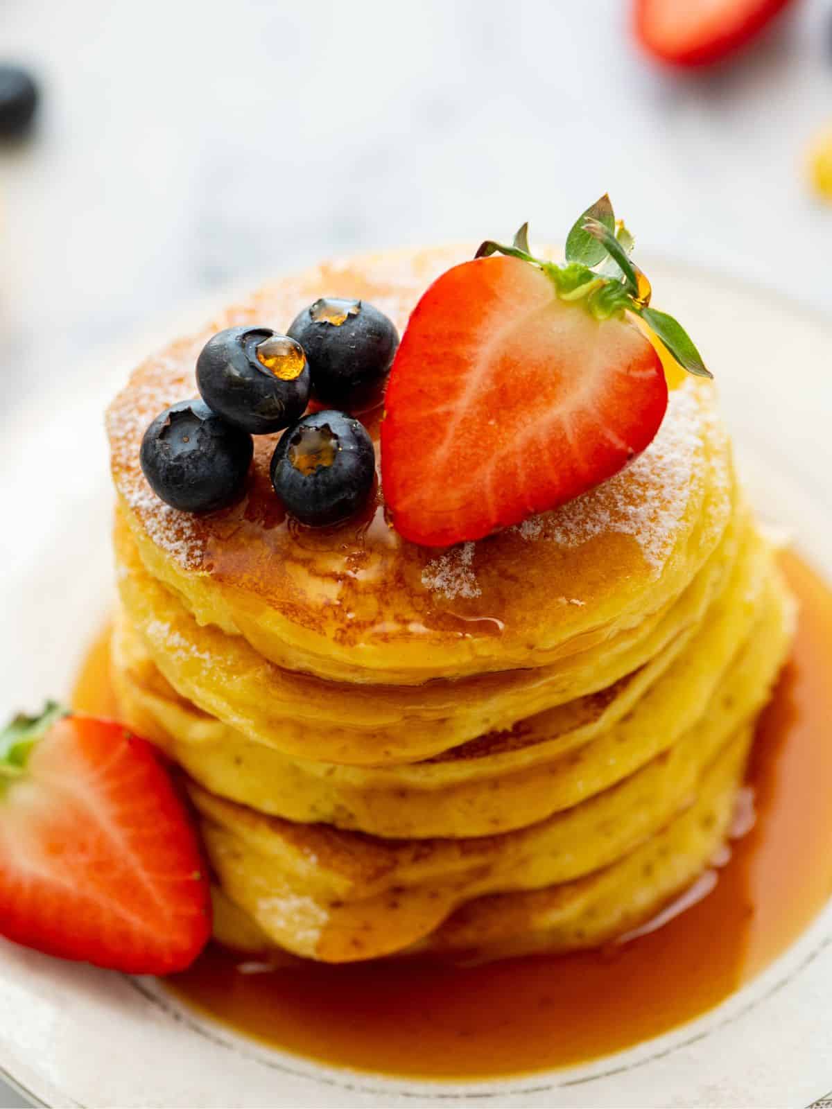 Stack of fluffy lemon ricotta pancakes on plate topped with maple syrup, blueberries, and strawberries.