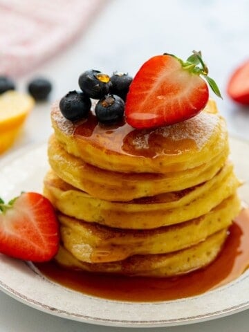 Stack of fluffy lemon ricotta pancakes on plate topped with maple syrup, blueberries, and strawberries.