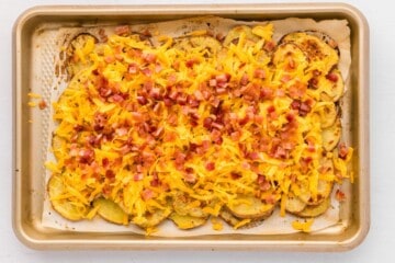 baked potato slices layered on a baking sheet topped with shredded cheddar and bacon.