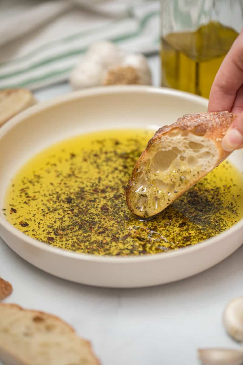 Toasted baguette being dipped into olive oil bread dip with fresh garlic in background.