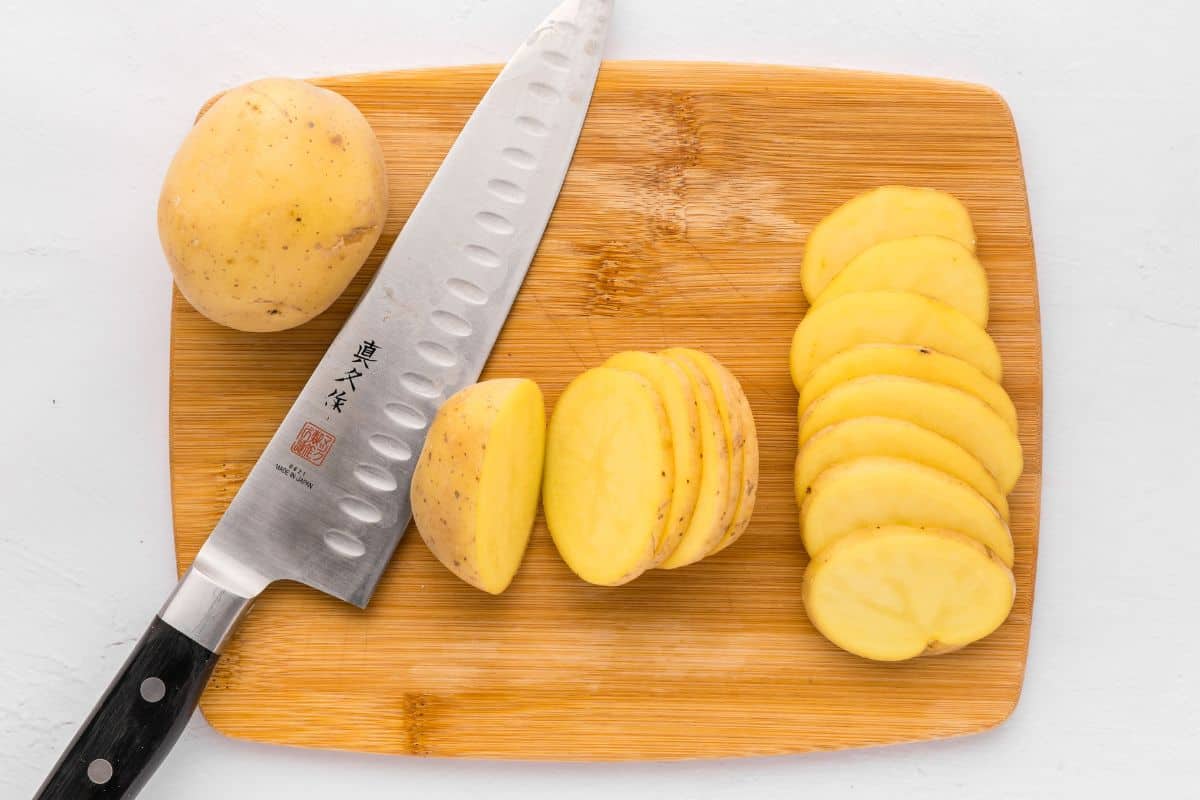 Yukon gold potatoes on wooden cutting board being cut into ¼-inch thick slices.