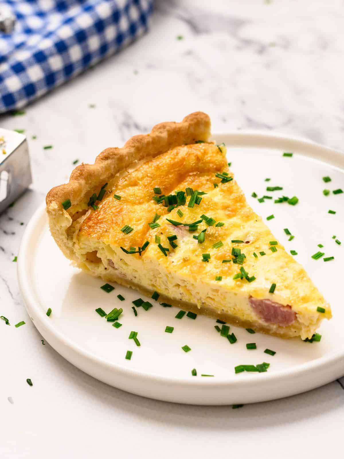 Slice of homemade quiche topped with chopped chives on white plate.
