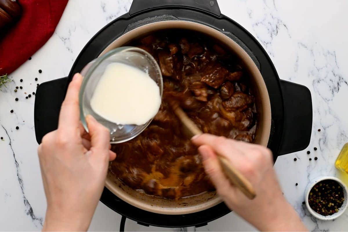 Pouring cornstarch slurry into inner pot with beef bourguignon while stirring with wooden spoon.