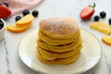 Stack of fluffy lemon ricotta pancakes on plate topped with a dusting of powdered sugar with lemon slices in background.
