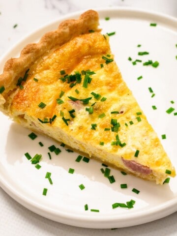 Slice of ham and cheese quiche topped with chopped chives on white plate.