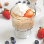 Tapioca pudding in small serving bowl topped with fresh whipped cream and strawberries with berries and cinnamon sticks in background.