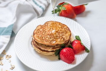 Cottage cheese pancakes stacked on white plate with syrup being poured over them next to fresh strawberries.