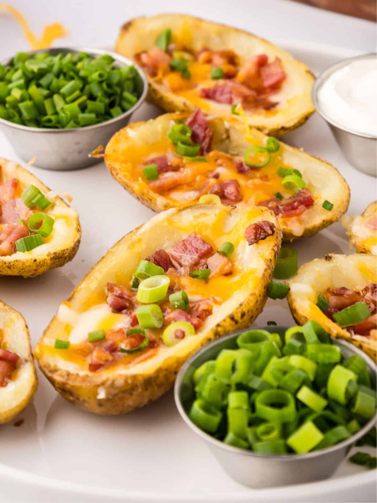 Crispy air fryer potato skins on white serving platter next to small bowls of green onions and sour cream.