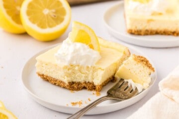 Slice of lemon cheesecake bars with fork taking bite out of bar topped with whipped cream and sliced lemon.