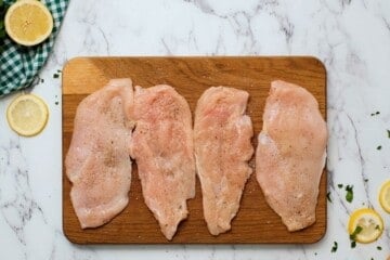 Thin chicken cutlets on wooden cutting board seasoned with salt and pepper and fresh lemon slices in background.