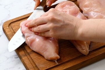 Knife cutting chicken breasts in half lengthwise to create cutlets.