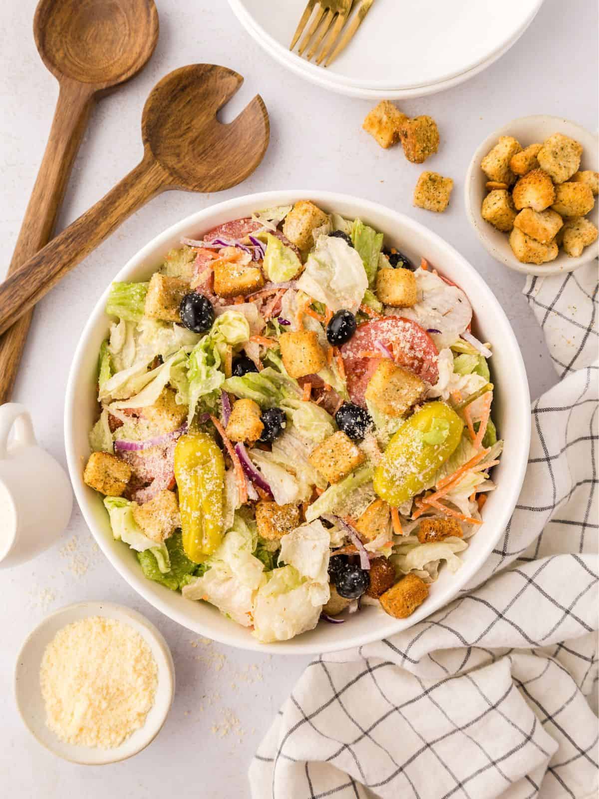Copycat Olive Garden salad made with iceberg lettuce mix, olives, tomatoes, and croutons in large white serving bowl next to small bowl of parmesan cheese.