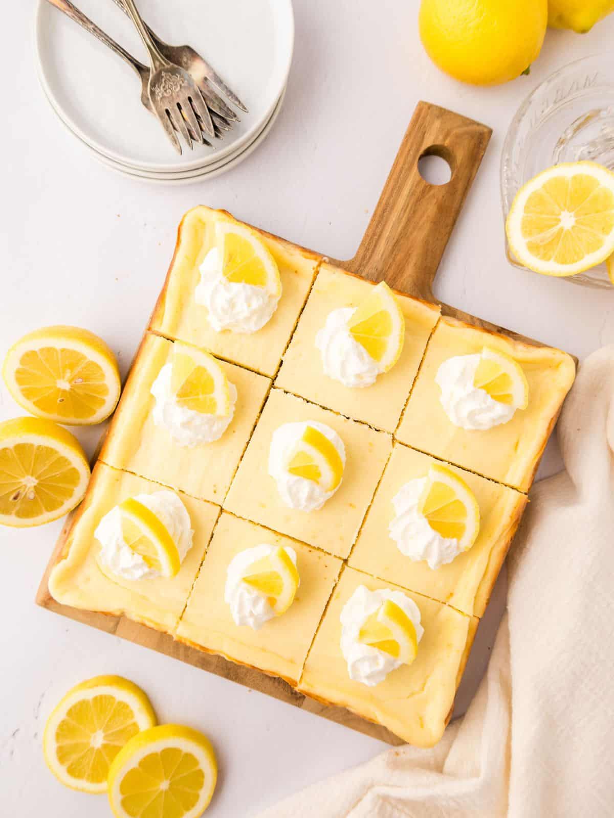 Baked Lemon Cheesecake Bars on wooden platter cut into 12 pieces and topped with dollops of whipped cream and lemon wedges.