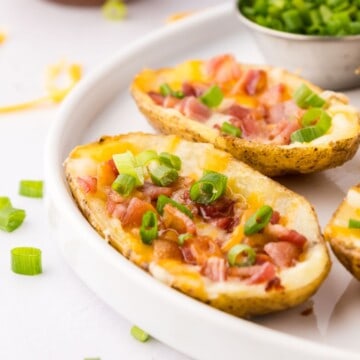 Crispy air fryer potato skins on white serving platter with small dishes of green onions, sour cream, and bacon for topping.