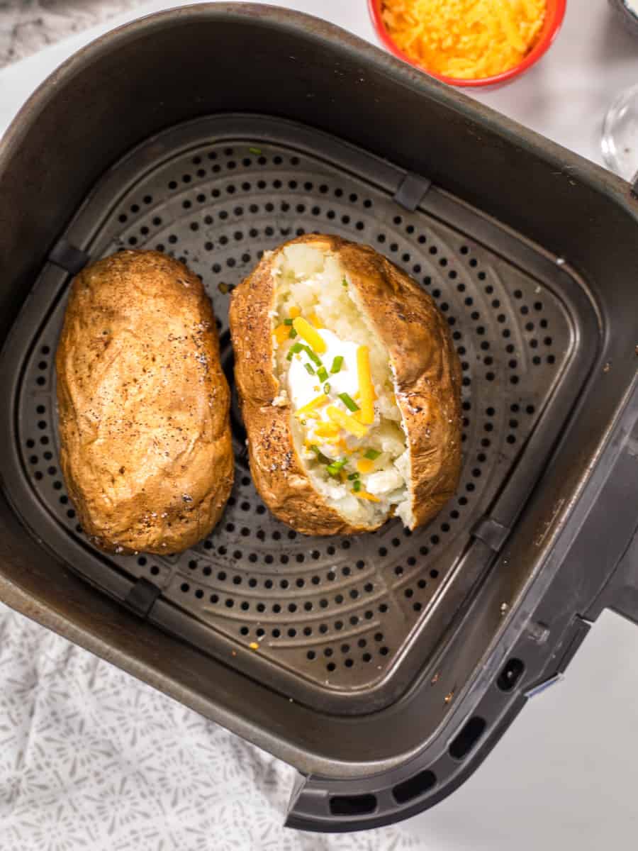 Two large baked potatoes in air fryer, one cut open and topped with butter, sour cream, and chives.
