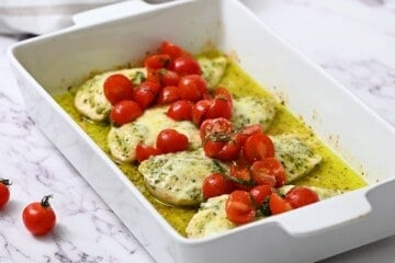 Baked pesto chicken in white baking dish topped with fresh cherry tomato and basil topping.