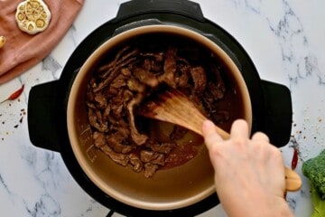 Wooden spoon scraping the bottom of the inner pot of a pressure cooker after adding the brown sauce and browning the beef.