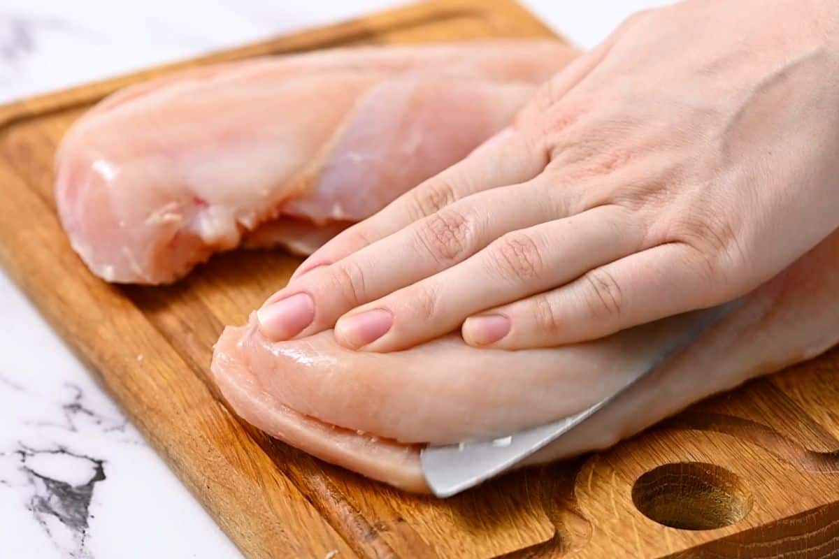 Knife cutting thick chicken breasts into chicken cutlets.