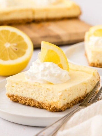 One lemon cheesecake bar on white plate topped with whipped cream and a lemon wedge.