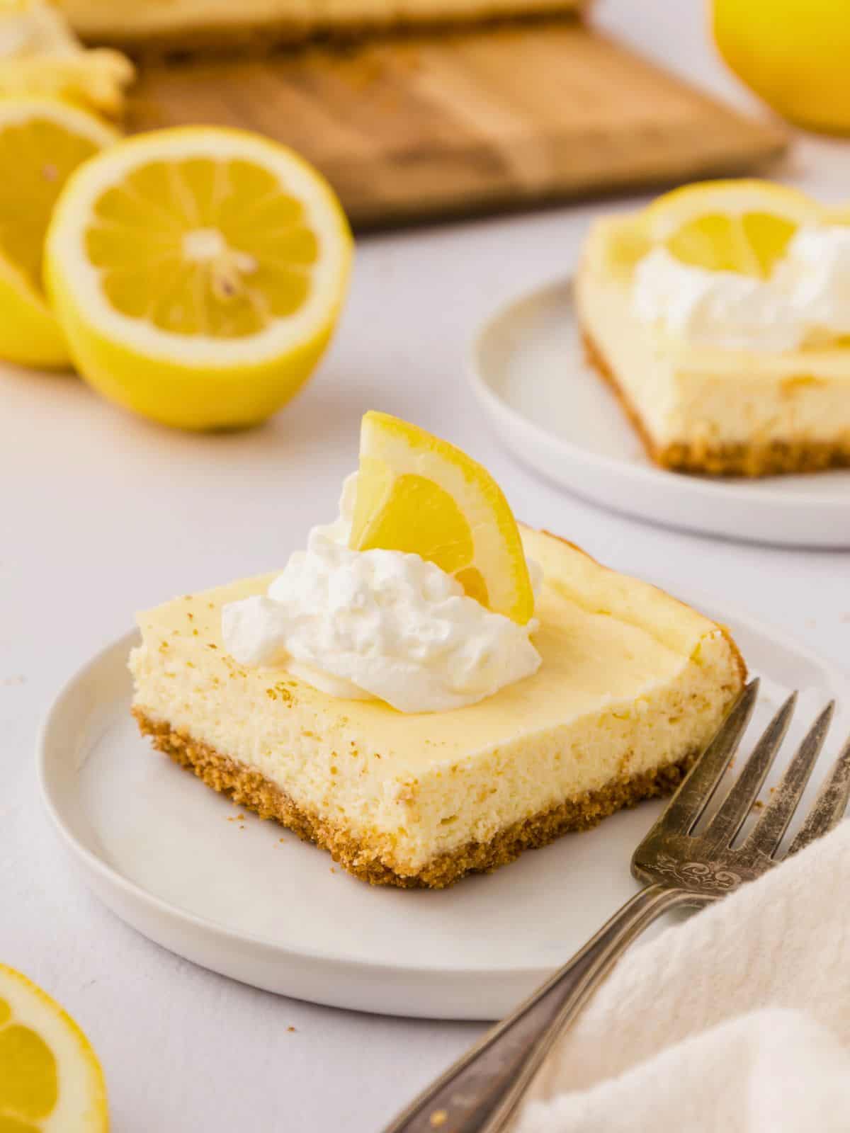 One lemon cheesecake bar on white plate topped with whipped cream and a lemon wedge.