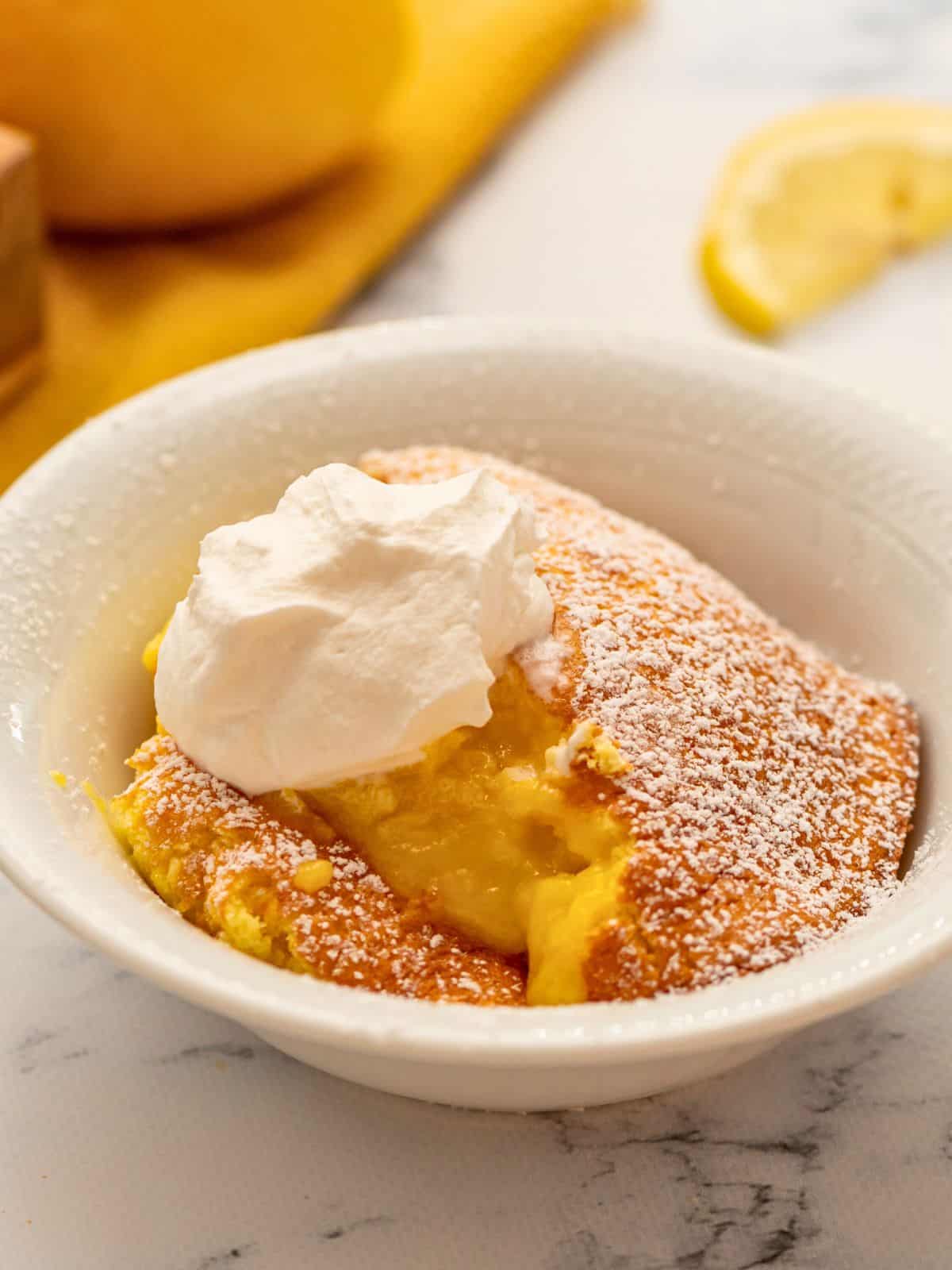 Baked Lemon Pudding Cake served in white dish topped with powdered sugar and whipped cream.