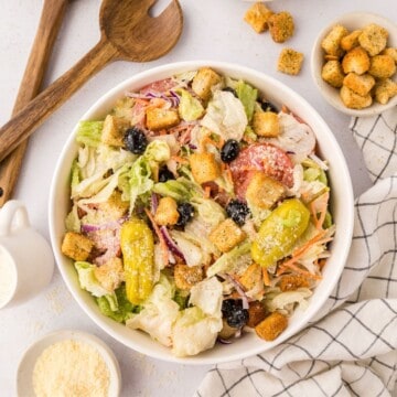 Copycat Olive Garden salad made with iceberg lettuce mix, olives, tomatoes, and croutons in large white serving bowl next to small bowl of parmesan cheese.