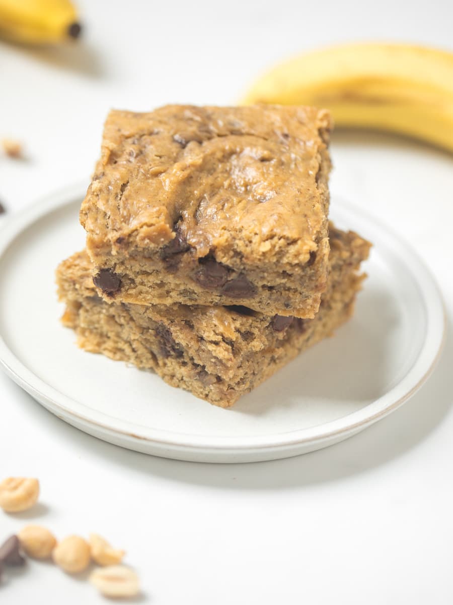Two slices of peanut butter banana bars on a white plate with peanuts and chocolate chips to the side.