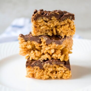 Three peanut butter Rice Krispie treats stacked on top of each other on white plate that are iced with chocolate.