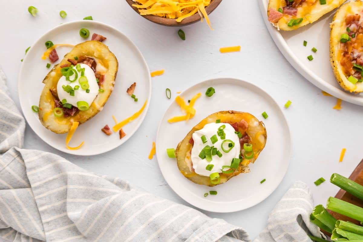 Crispy air fryer potato skins on white plates topped with green onions, sour cream, and bacon and green onions in background.