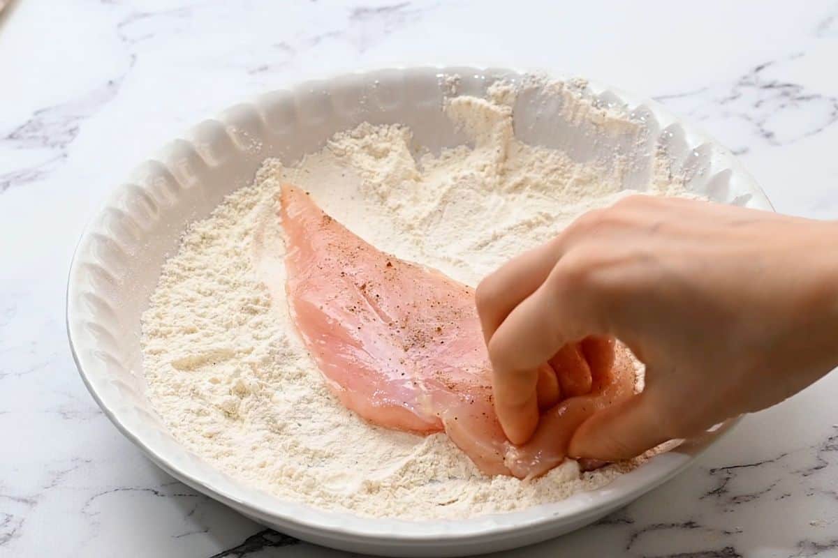 Chicken cutlet being coated in seasoned flour mixture.