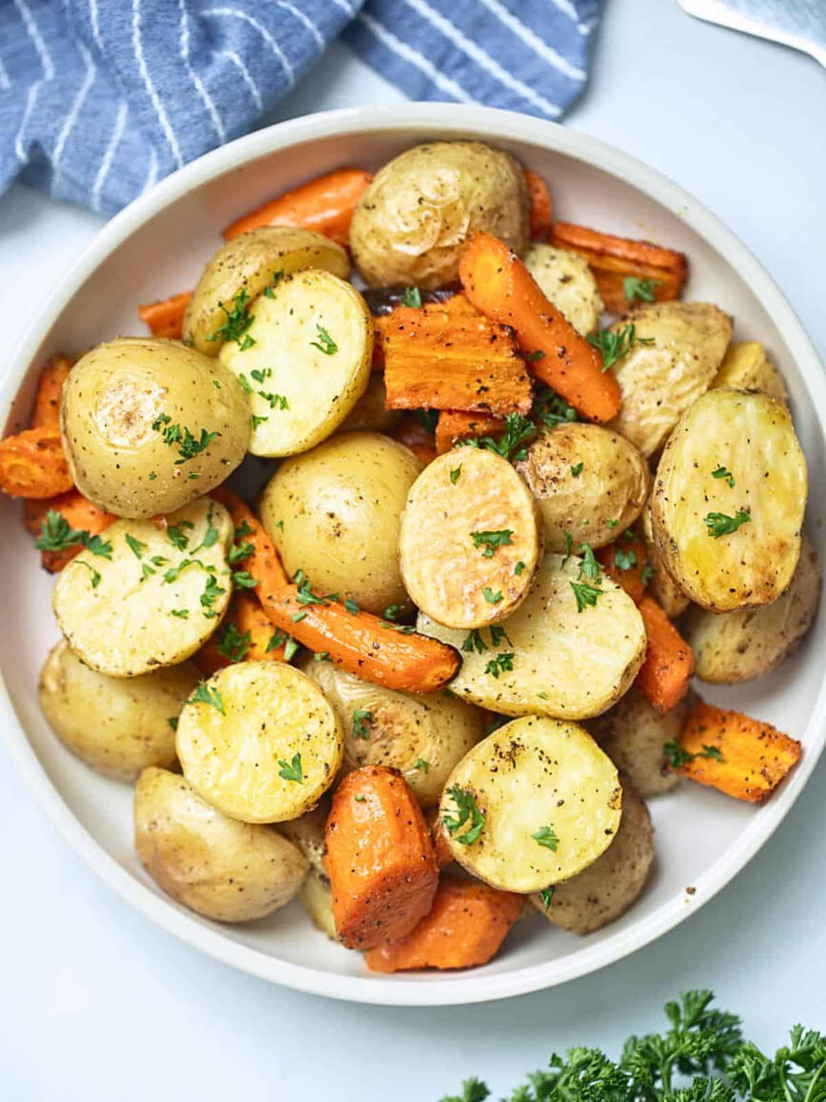 Roasted potatoes and carrots in white serving bowl next to fresh parsley.