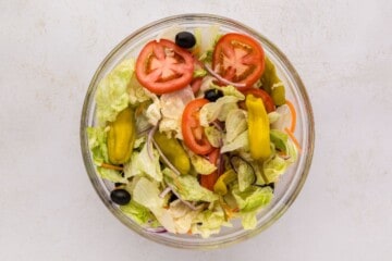 Clear mixing bowl with iceberg, cabbage, onions, carrots, olives, tomatoes, and pepperoncini peppers tossed together.