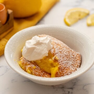Baked Lemon Pudding in white bowl topped with whipped cream and powdered sugar.