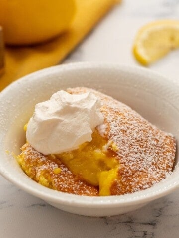 Baked Lemon Pudding in white bowl topped with whipped cream and powdered sugar.