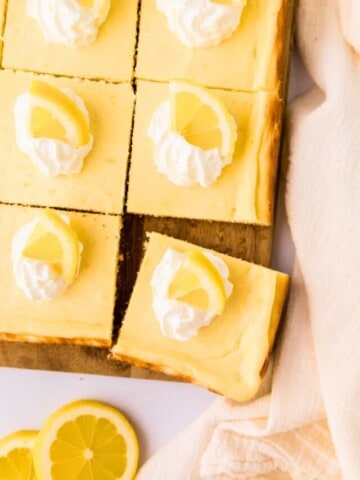 Baked Lemon Cheesecake Bars on wooden platter cut into 12 pieces and topped with dollops of whipped cream and lemon wedges.