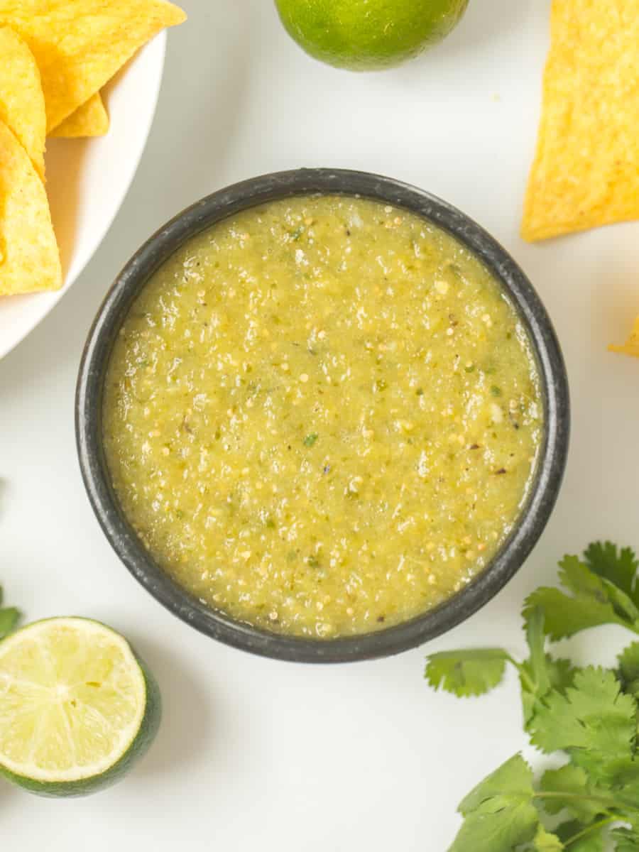 Homemade Salsa Verde in black bowl with corn tortilla chips on the side with cilantro and limes in the background.