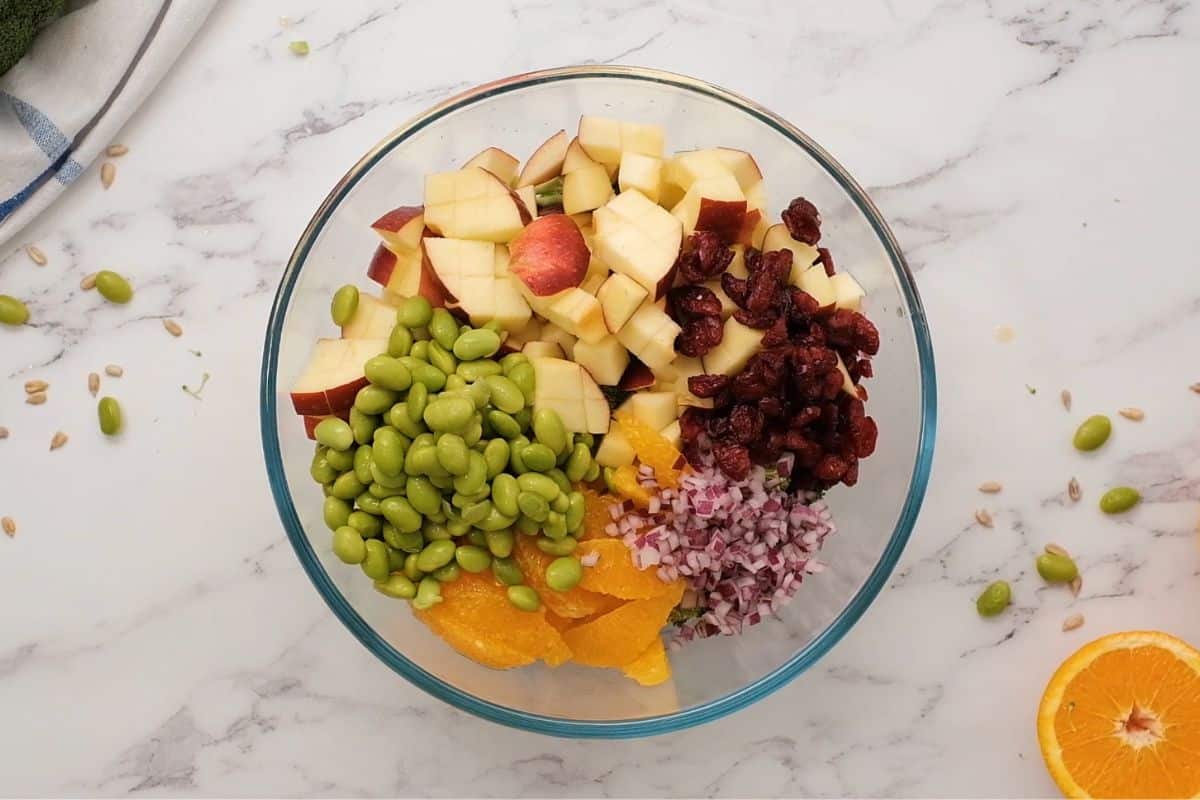 Broccoli, apples, edamame, oranges, onions, and cranberries in bowl.