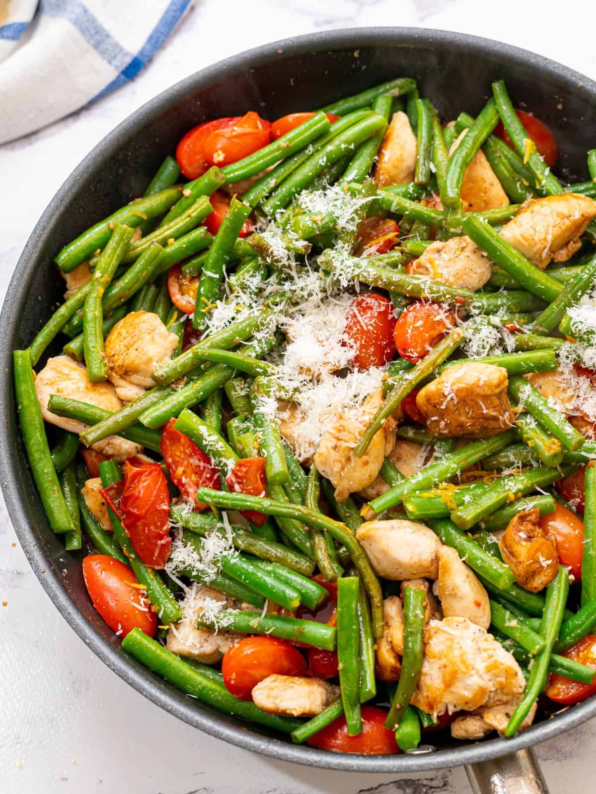 Large skillet with chunks of chicken, green beans, cherry tomatoes, and parmesan.