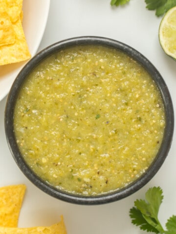 Homemade Green Salsa Verde in black bowl with corn tortilla chips on the side with cilantro and limes in the background.