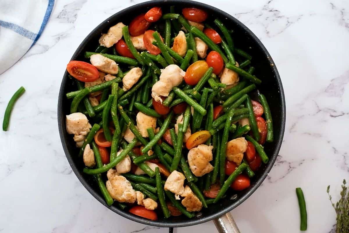 Green beans and tomatoes added to browned chicken in skillet with fresh thyme in background.