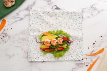 Tea towel with spring roll wrapper on it topped with lettuce, peppers, mango, carrots, mint, and shrimp before rolling into spring roll.