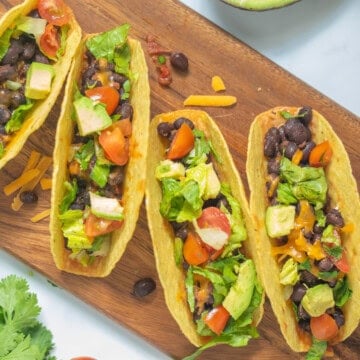 Black Bean Tacos on wooden cutting board topped with fresh tomatoes, lettuce, cilantro, and avocado.