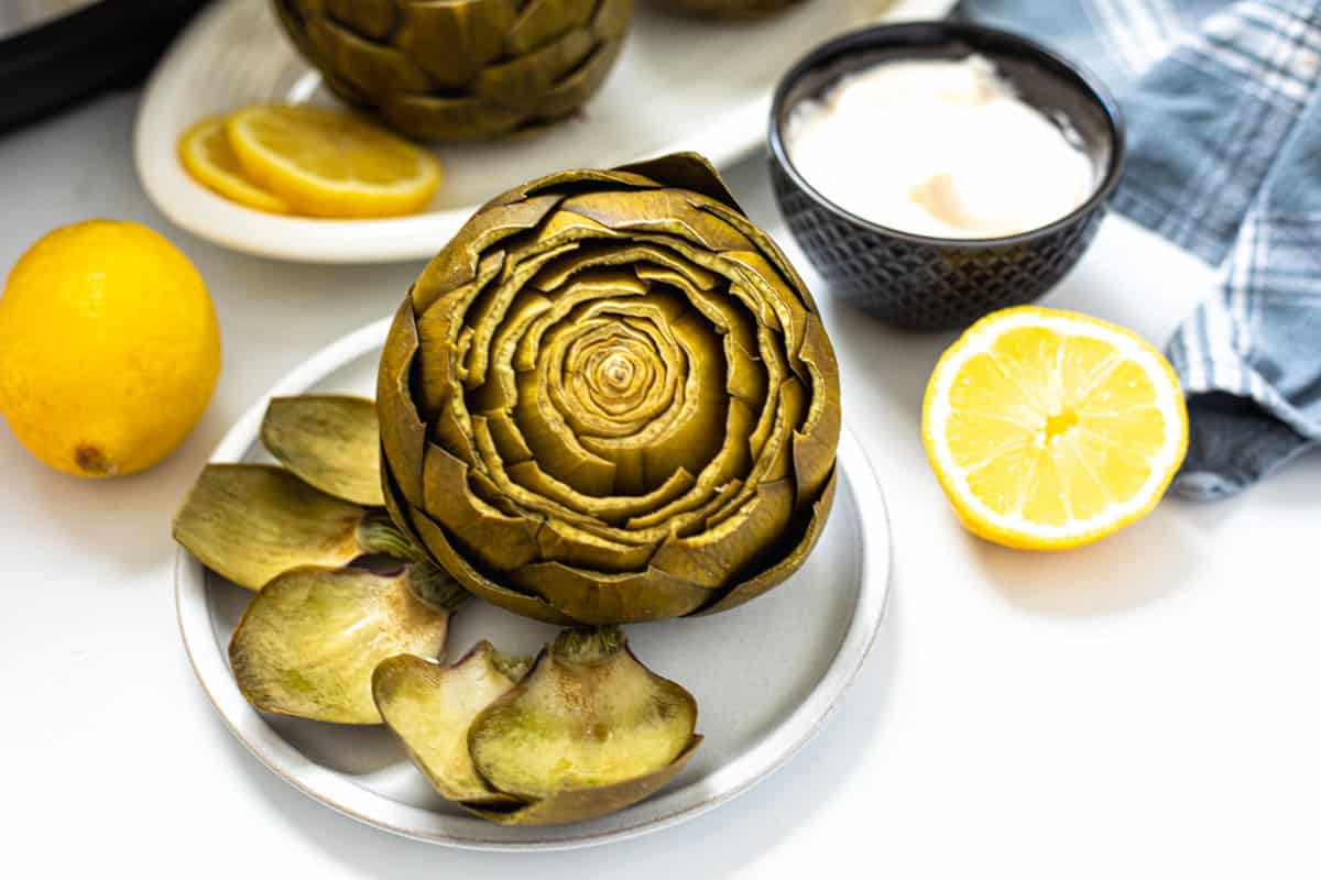Steamed whole artichoke on white plate with Instant Pot and lemon in background.