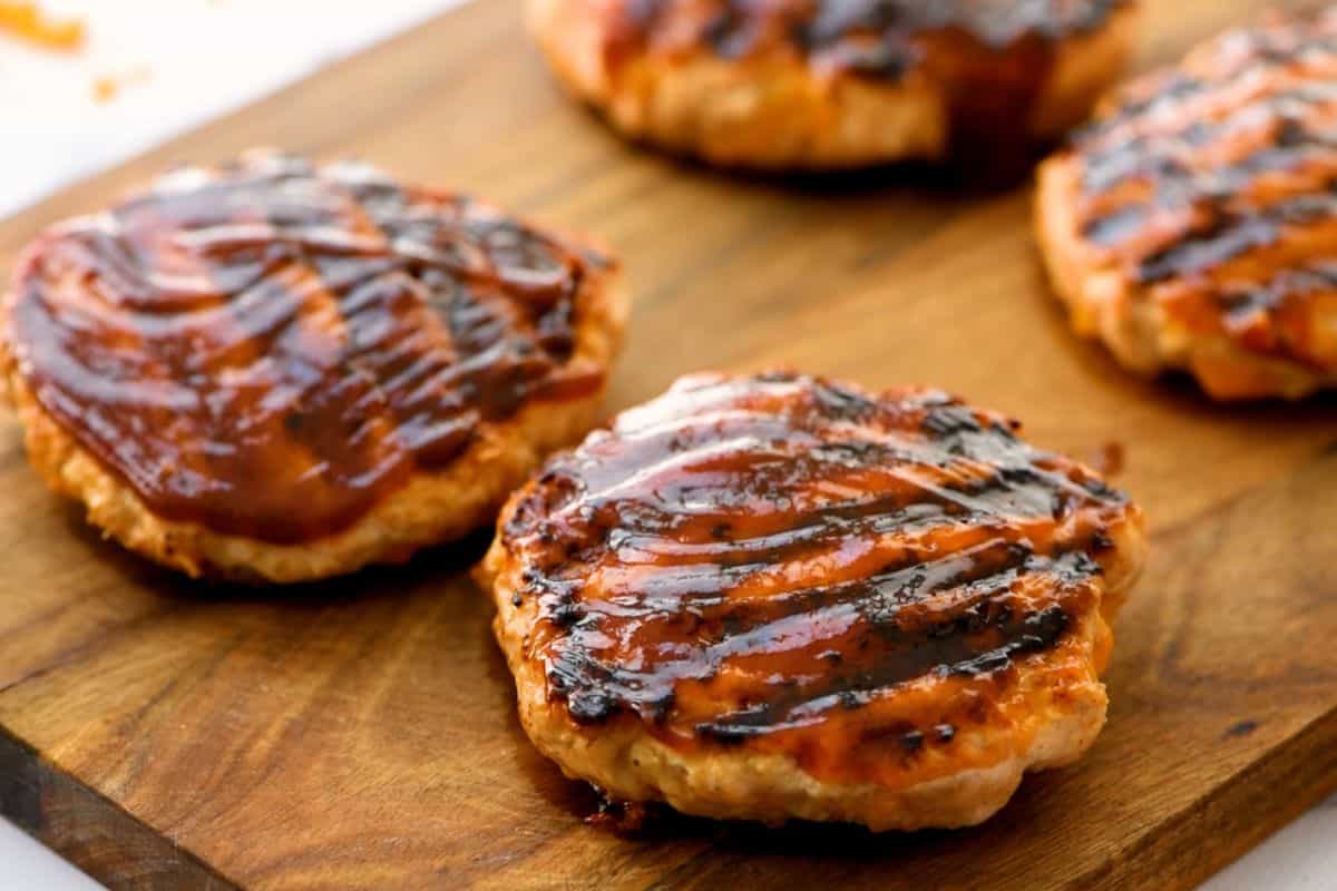 Four chicken burgers on wooden platter basted in BBQ sauce.