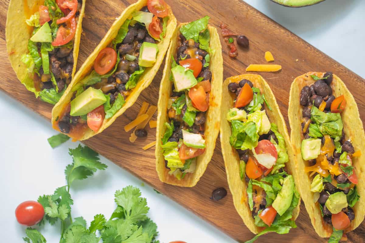 Black Bean Tacos on wooden cutting board topped with fresh tomatoes, lettuce, cilantro, and avocado.