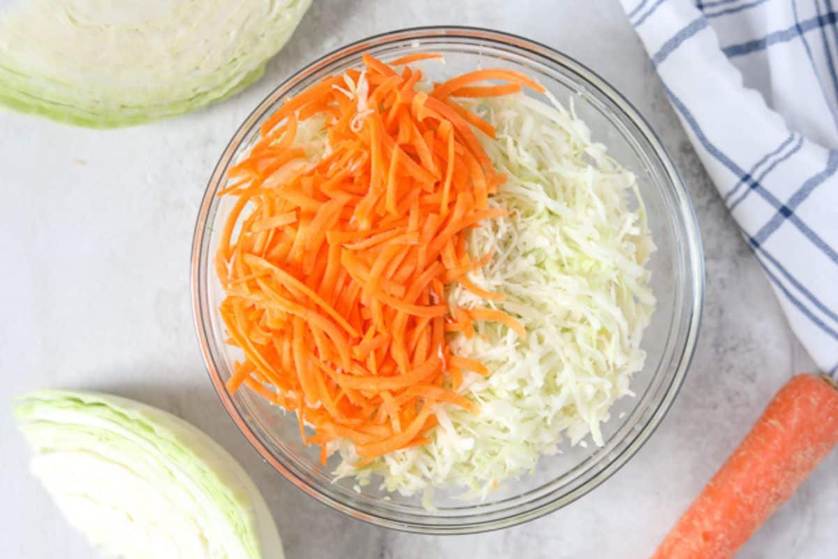 Clear mixing bowl with shredded green cabbage and shredded carrots.