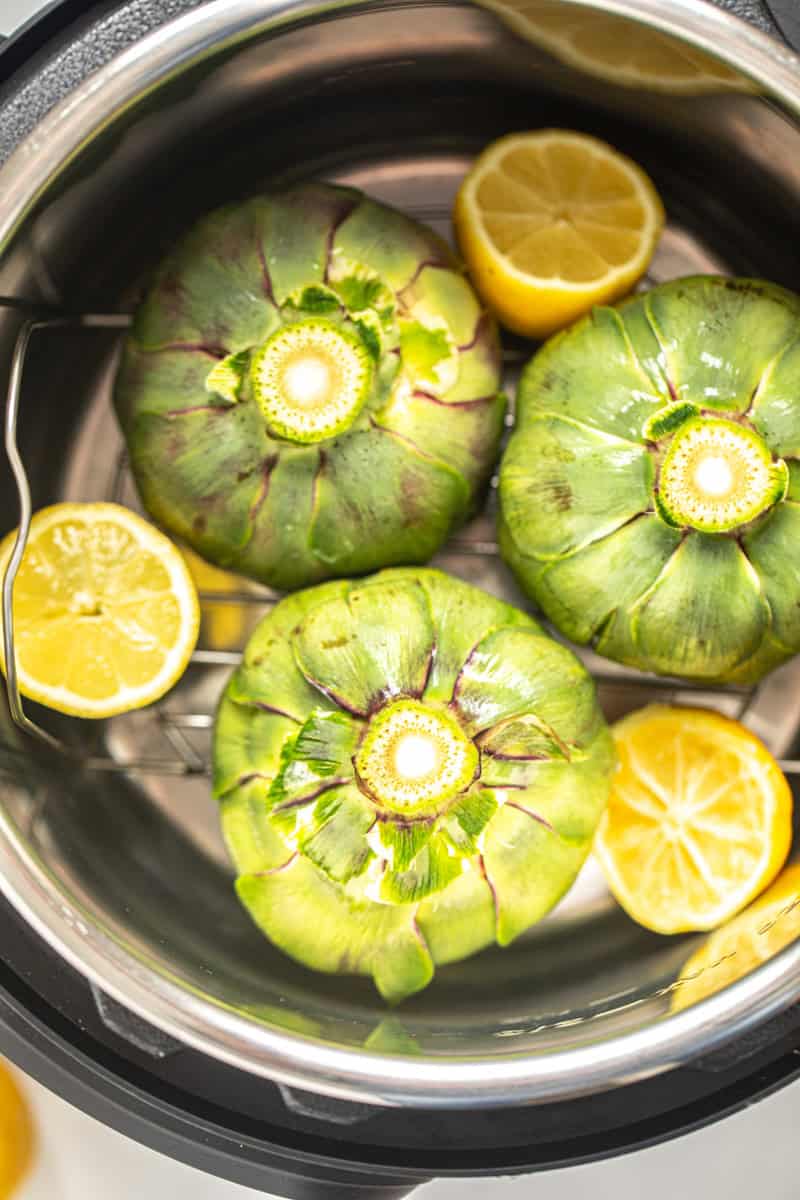 Three cleaned whole artichokes inside inner pot on metal trivet with sliced lemon.